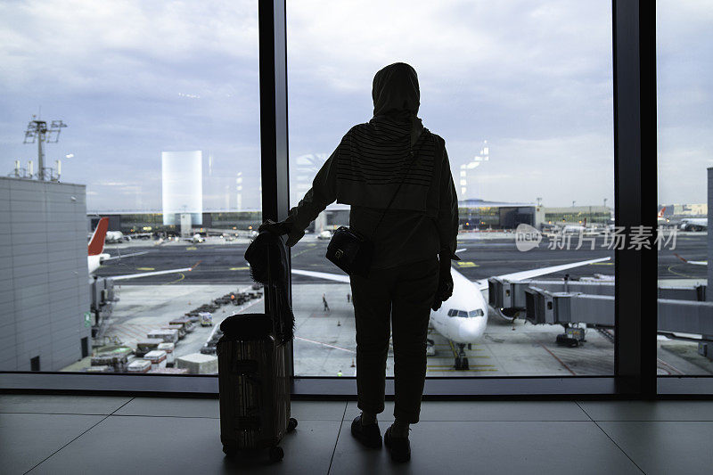旅客拿着行李站在机场窗口看日落。一名女子在候机室看着飞机，在登机门等待起飞。