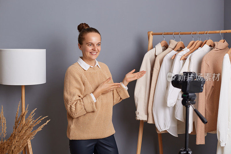 可爱乐观的女性造型师或设计师穿着米色毛衣，展示她的新系列，为vlog拍摄视频，站在灰色的墙壁上，衣服挂在衣柜的架子上。