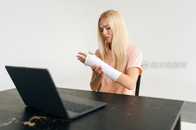 沮丧的金发年轻女子的肖像骨折的手臂包裹在石膏绷带有视频通话，使用笔记本电脑在线咨询。