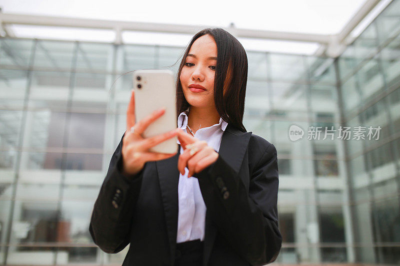 亚洲年轻美丽的女商人看着她的手机，在商业中心前笑