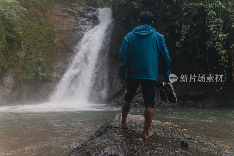 一名亚洲男子在热带雨林中拿着鞋子欣赏瀑布美景