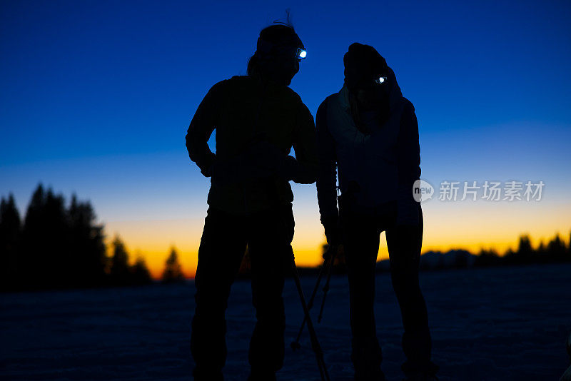 两名女子滑雪者，额头顶着手电筒，站在雪坡上