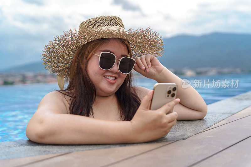 超重的年轻女子橙色swimsuitÂ在游泳池里放松地拿着智能手机。肥胖女人度假旅行用手使用手机