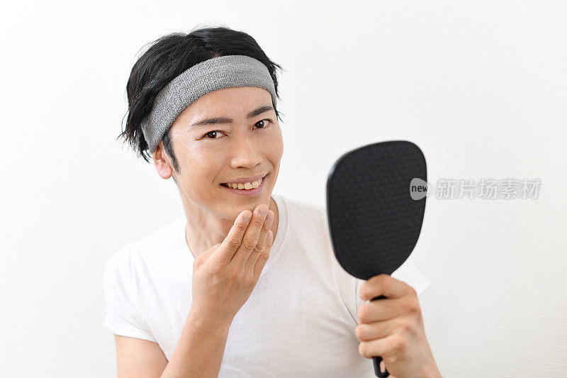 一位年轻的亚洲男子正微笑着看着手镜，注意着自己的皮肤护理。
