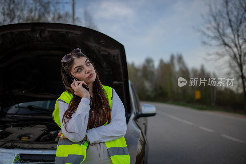 年轻的压力很大的女子站在她抛锚的汽车旁边，用手机求救