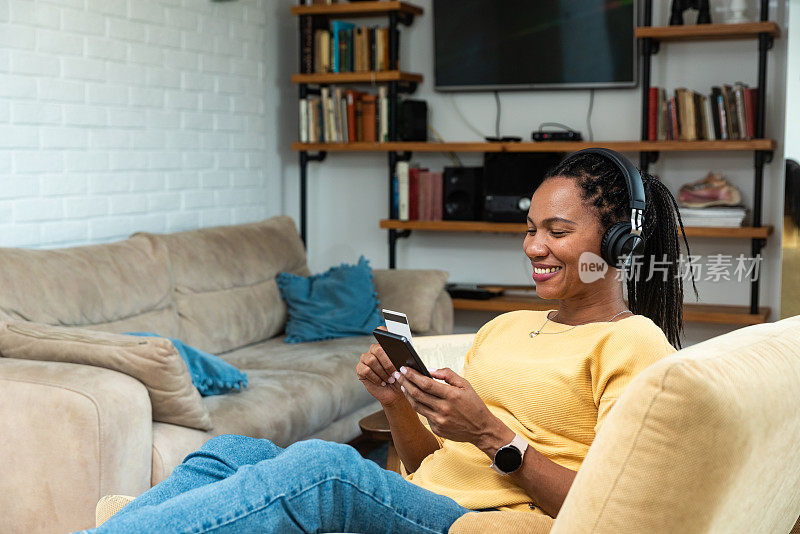 年轻微笑的非洲裔美国妇女拿着信用卡使用智能手机。网上支付、家居购物、电子商务、网上银行、消费理财、电子商店概念自椅。