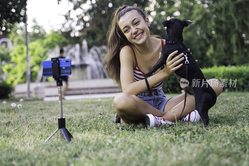 美丽的少女微笑着用手机在公园里拍宠物狗视频。网红录制视频。
