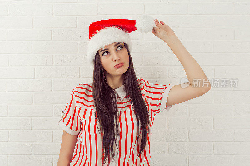 滑稽的女人戴着红色圣诞老人的帽子抬头看着绒球，颠覆了孤立的白砖墙背景。圣诞节的女人。