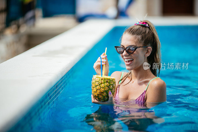 美丽的年轻女子在游泳池喝菠萝鸡尾酒