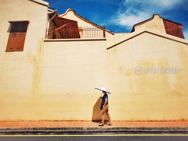 手机摄像头拍摄的是一名亚洲华人女士在马六甲镇的中午撑着伞走在街上的画面