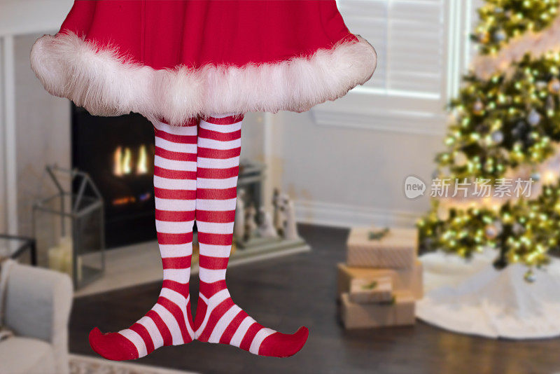 可爱的圣诞小精灵小女孩穿着条纹长袜站在装饰好的圣诞客厅里，背景是礼物和圣诞树
