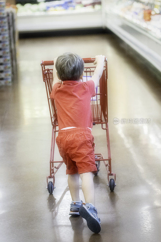 一个三岁的小男孩在超市的过道里推着一个小购物车