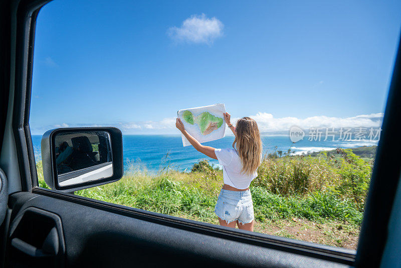 一名年轻女子在沿海公路上的车外看地图问路。夏威夷的山海景观