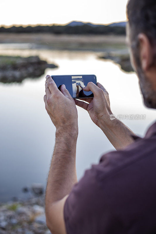 西班牙马德里，加拉帕加尔，一名中年男子在瓦尔马约尔水库的一个土堆顶上用智能手机拍照