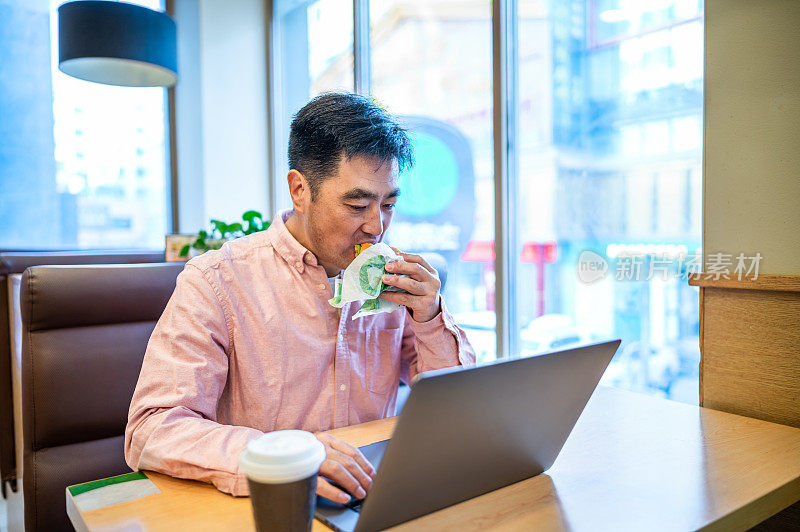 亚洲中年男子在咖啡店吃汉堡用笔记本电脑