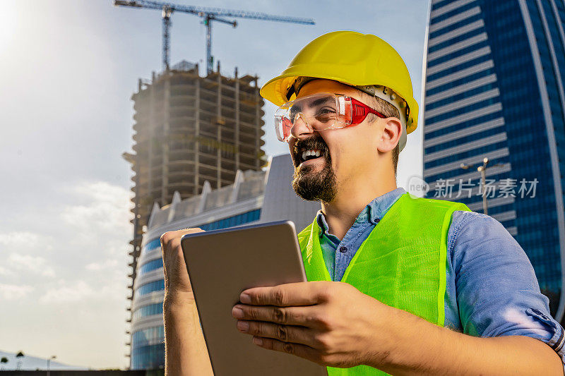 快乐的年轻工程师或建筑师，穿着防护服，在现代商业建筑和施工现场前使用数码平板电脑