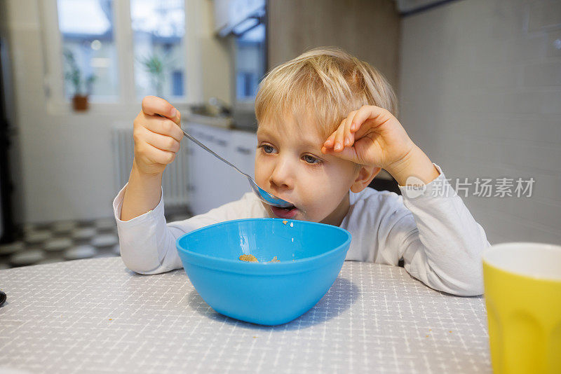 金发蹒跚学步的孩子坐在餐桌上，从碗里吃麦片，看起来很累
