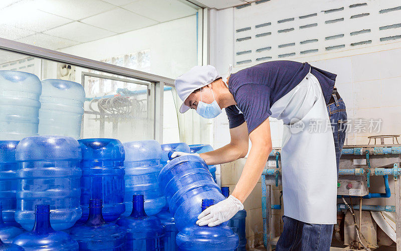 在饮水厂工作的男子工人穿着工作服，脸上戴着防护口罩，在装运前检查蓝色水加仑。