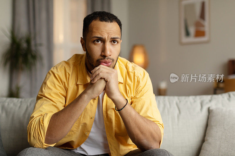 黑人坐在沙发上，表情若有所思，在家里感到悲伤，遇到问题，承受着压力