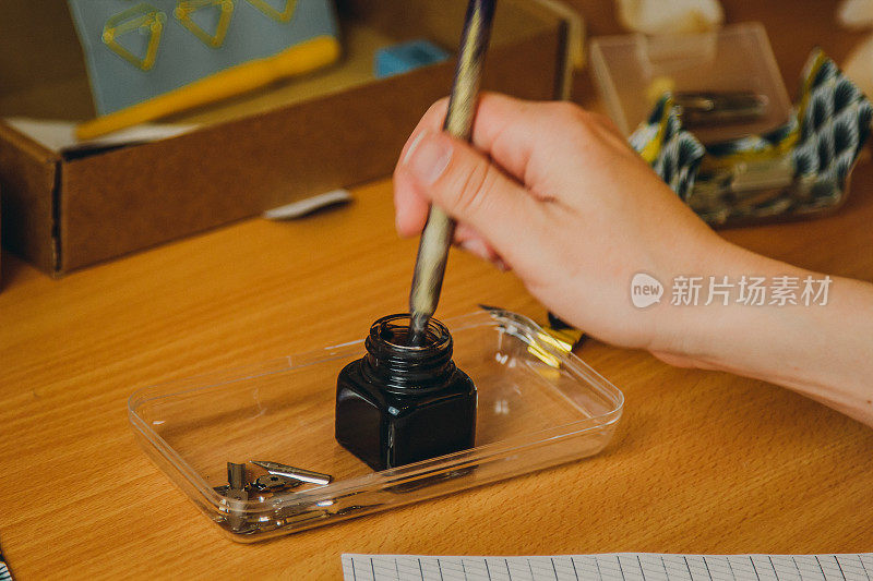 女性用手将墨笔浸入墨水瓶。木质书桌上的文具近距离摆放。拼写课和书法练习