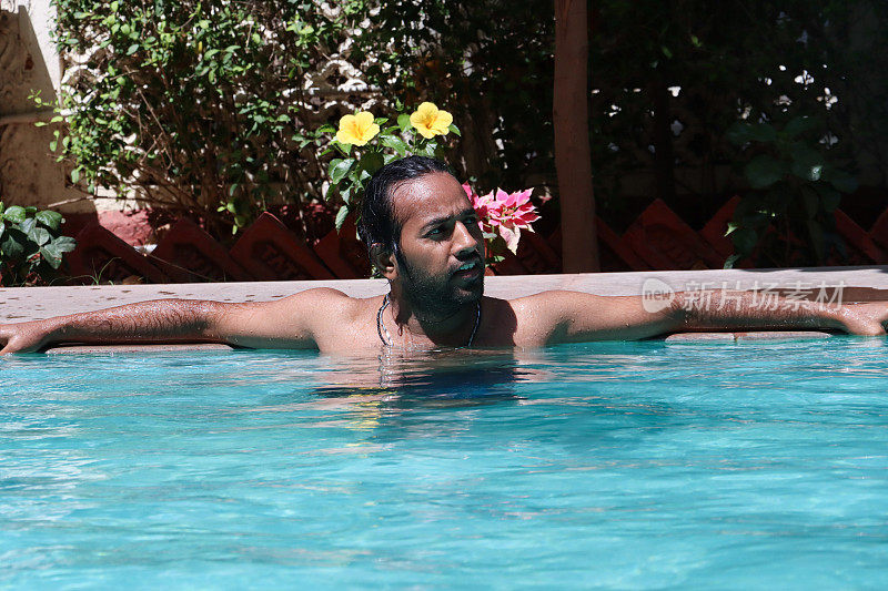 印度男子站在肩部深的清澈的水中，倚靠在室外，游泳池，绿松石蓝色马赛克瓷砖，手臂沿着墙壁伸展，波涛起伏的水面反射，重点放在前景