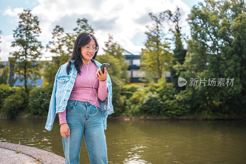 戴眼镜的亚洲女游客在河边用智能手机听音乐