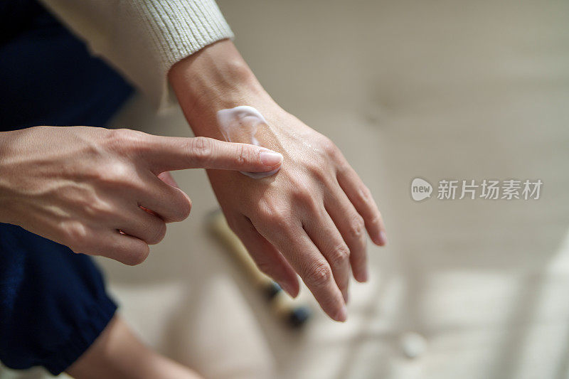 在这张剪短的图片中，一位中年亚洲女性正在用润肤霜涂抹双手干燥的皮肤，阳光温柔地照在她的皮肤上，突显出滋养的姿态。