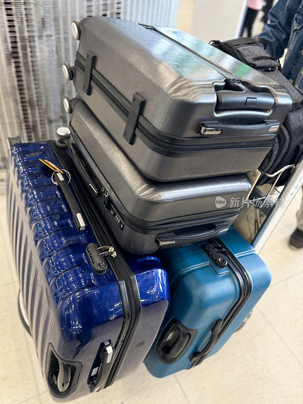 特写照片中，无法辨认的乘客穿着牛仔夹克，操纵着一堆轮式行李箱的行李车，在支柱和可伸缩皮带旁排队，机场出发区，闪亮的瓷砖地板，重点放在前景上