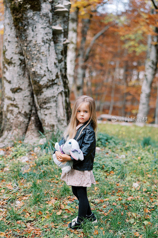 小女孩手里拿着一只玩具兔子，半转着身子站在秋天的森林里
