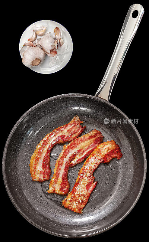 油炸美食多汁脆培根片在大型重型煎锅与一盘大蒜鳞茎和丁香设置在直接观看上方孤立的黑色背景高分辨率库存图像