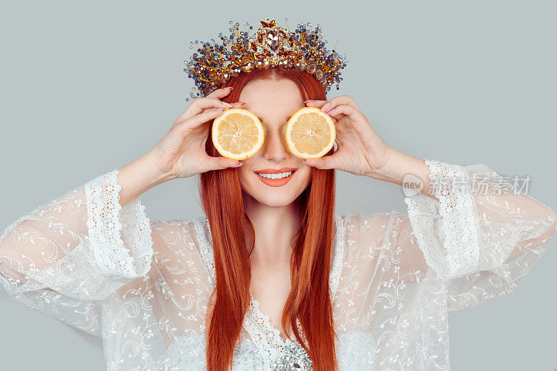 维生素C和美容。一个年轻的女人拿着覆盖眼睛的两片橙色柠檬作为一个眼睛区域皮肤面膜漂亮的女人与水晶皇冠上的头孤立在浅灰色的背景