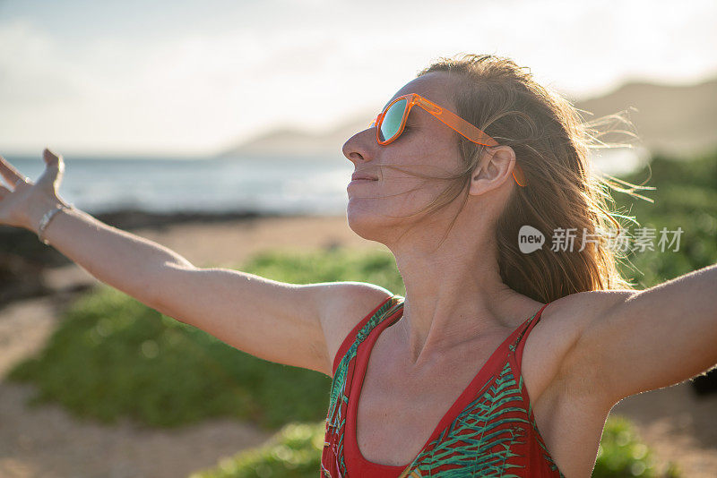 夕阳西下，拥抱大自然的快乐年轻女子;女人站在沙滩上，伸开双臂。