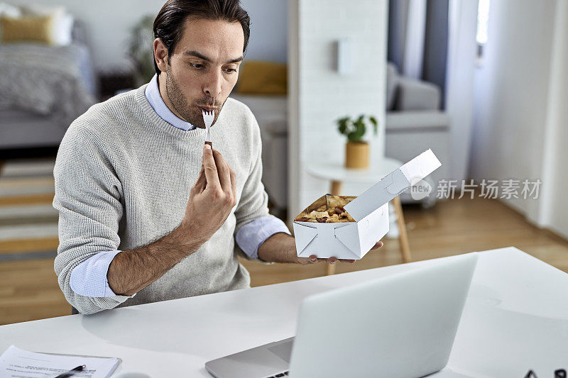 年轻的企业家使用笔记本电脑，在家工作时吃东西。