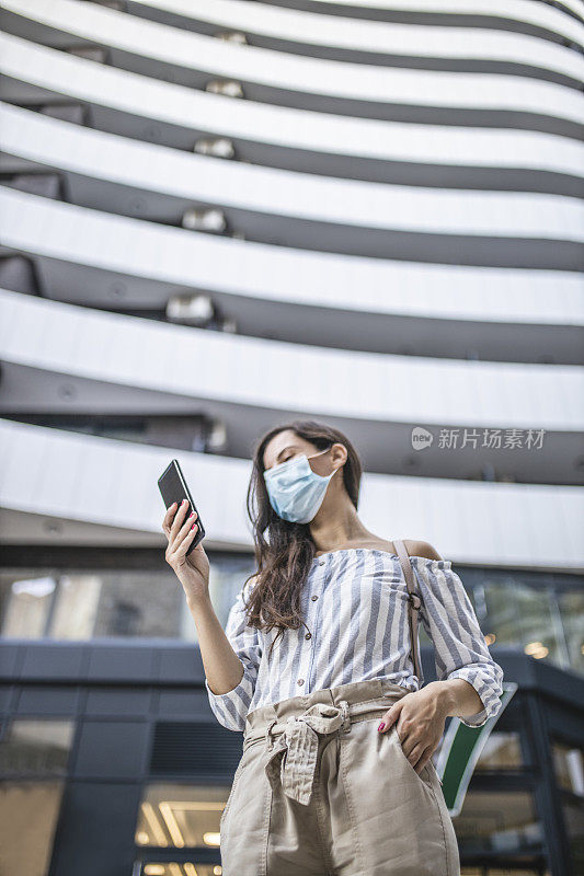 年轻女子站在城市街道上使用智能手机，脸上戴着防护面具。病毒大流行与污染概念。