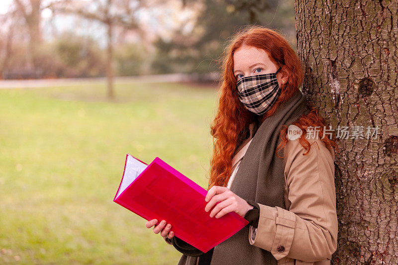 一个红头发的十几岁的学生靠在一棵树上在公园里学习她的课本