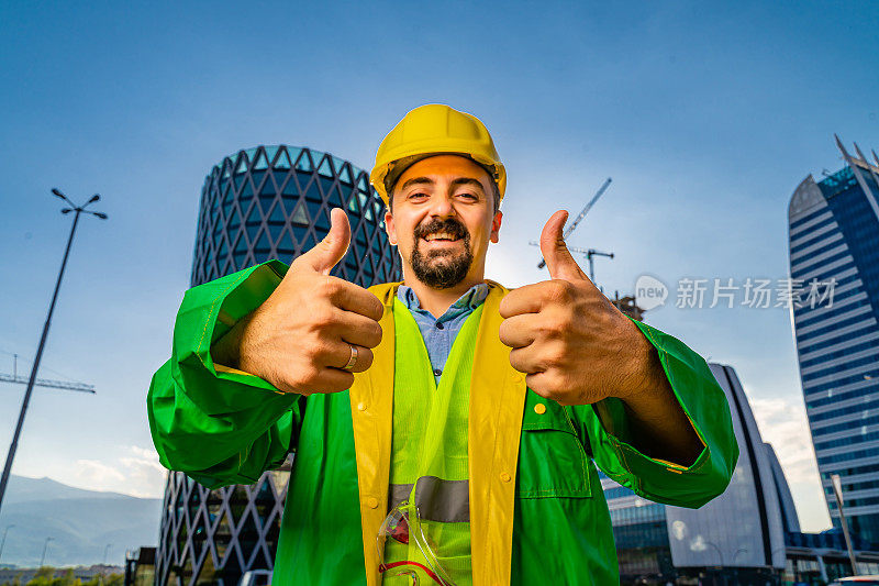 年轻的工程师戴着黄色安全帽，穿着绿色防护服，看着摄像机，在现代化的商业建筑和建筑工地前竖起大拇指。镜头光晕。低角度的观点