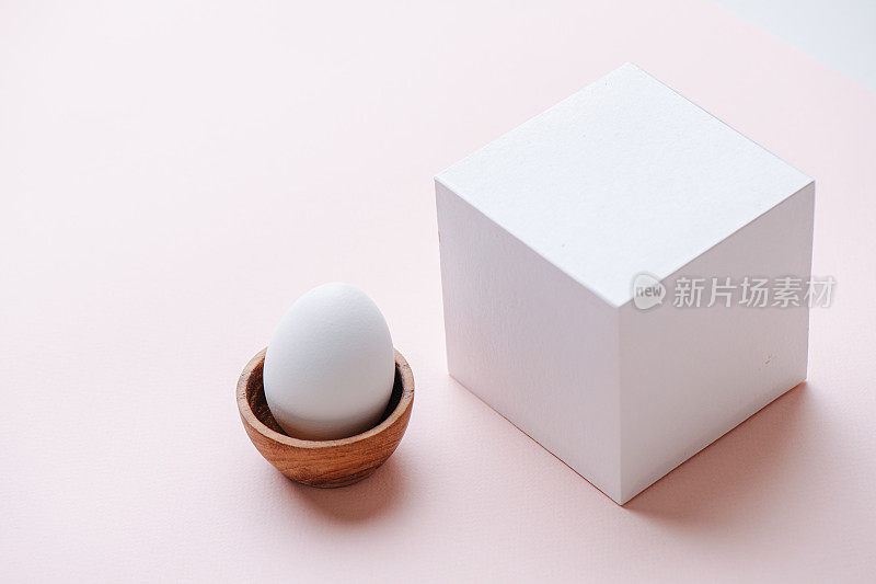 木蛋里的白蛋立在粉红色的表面上，旁边是一个白色的纸立方体