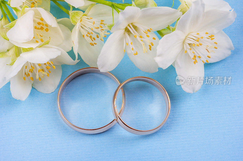 一对金色的结婚戒指与白色的茉莉花在一个蓝色的背景接近