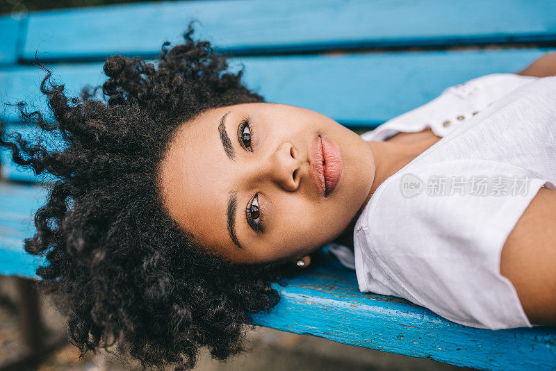 美丽的非裔美国女人正在公园的长椅上休息，看着镜头。美丽年轻的黑皮肤女性穿着白色t恤享受健康的生活。人,生活方式的概念