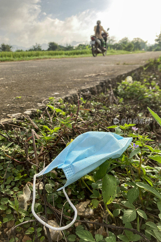 丢弃的脏兮兮的防护口罩躺在路边，一个骑摩托车的女士从后面经过。