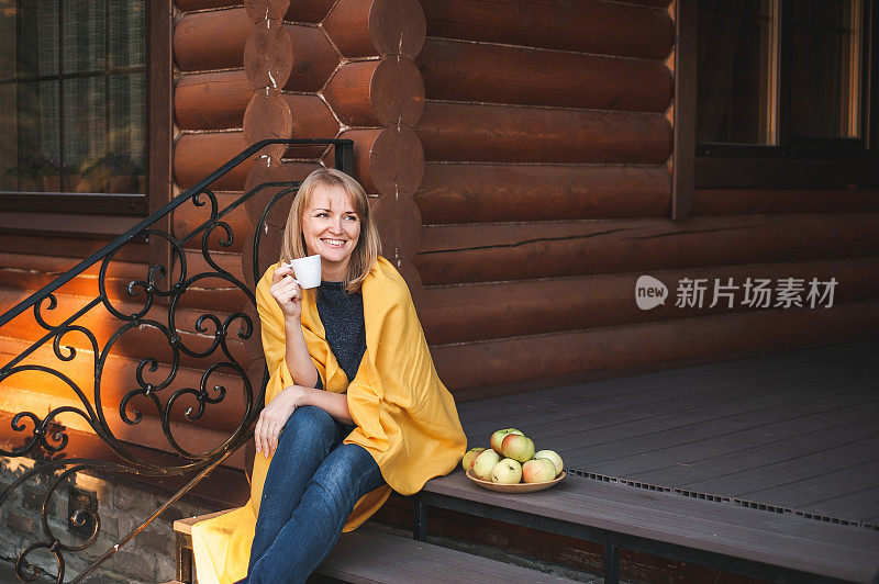 一位40岁左右的金发美女面带微笑，坐在木屋的门廊上，手里端着一杯咖啡，盘子里放着成熟的苹果。这个女人放松和享受乡村的秋天生活。