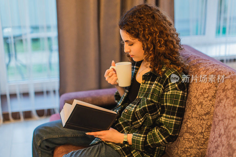 红发卷发的女孩坐在卧室的床上看书。休息在家里。一个女人拿着一个装着茶的白色杯子