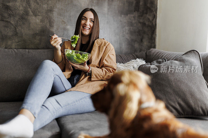 一个女人在家享受休息，吃着健康的蔬菜沙拉。她的狗也希望得到一些。