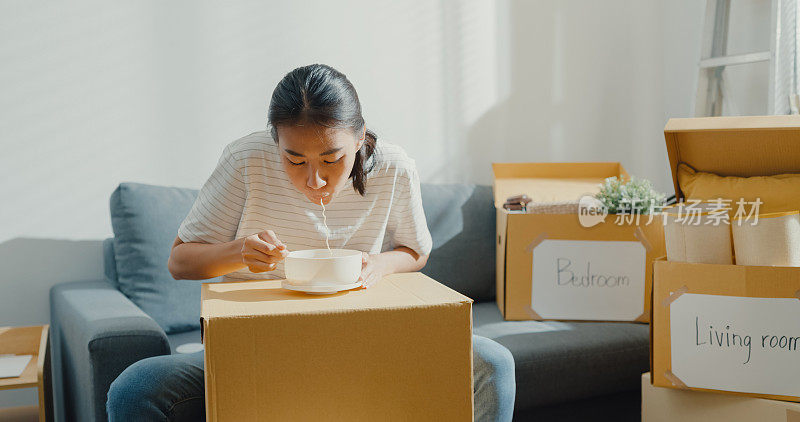 年轻的亚洲女人搬家坐在沙发上吃方便面后拆包。