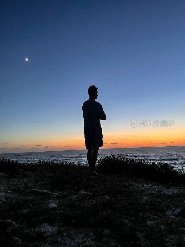 日落时，一个男人站在山上，背对着天空看海的剪影