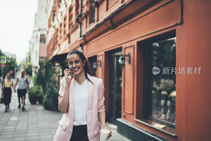 快乐的西班牙女性在城市城市化中行走，在漫游中积极的智能手机对话中微笑，真诚的时髦女孩戴着经典眼镜，在咖啡时间享受友好的电话