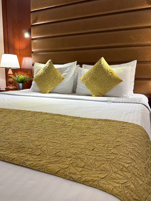 酒店房间双人床的特写图像，白色的酒店枕头靠在人造革床头板上，白色的羽绒被盖，金色的靠垫，在床头柜上照明的电灯，焦点在前景