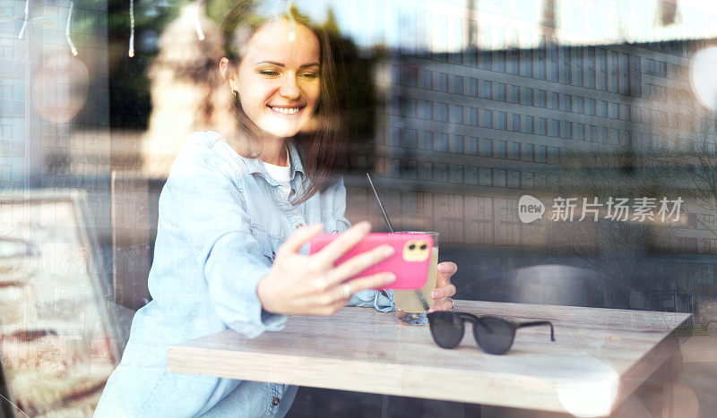 一名女子坐在咖啡馆里用智能手机自拍，城市街道的倒影映在咖啡馆的窗户上。