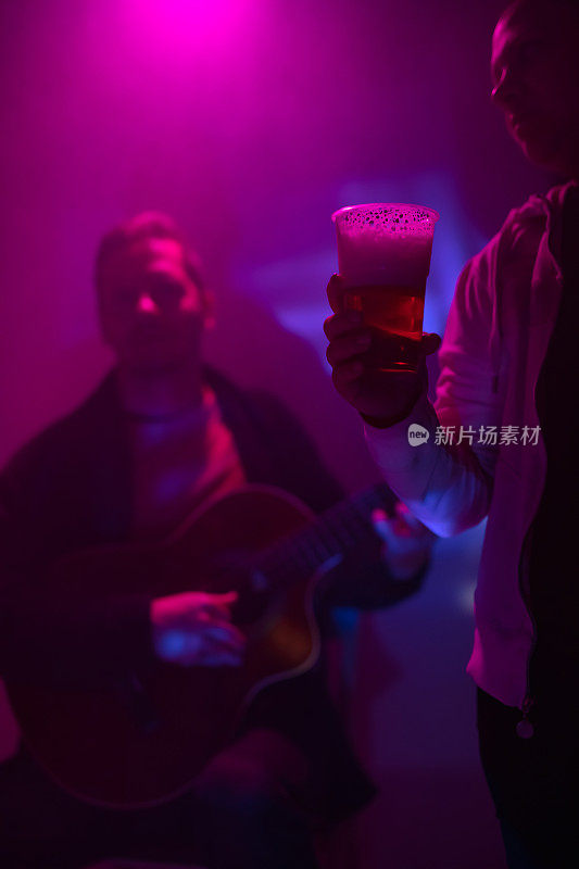 一名女子拿着一个塑料啤酒杯，在俱乐部背景中有一位音乐家在弹吉他。