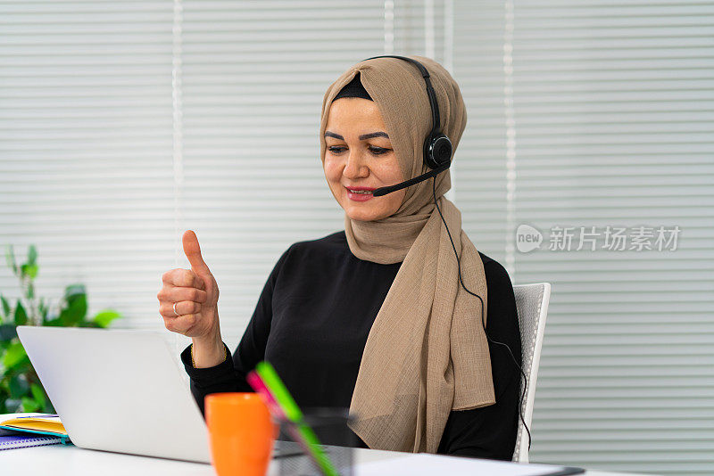 穆斯林妇女戴着耳机看着笔记本电脑在办公室通过视频通话